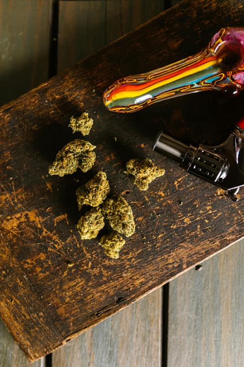Marijuana Buds with Pipe and Lighter on Wooden Surface Dried cannabis buds placed on a rustic wooden board next to a colorful glass pipe and a lighter.